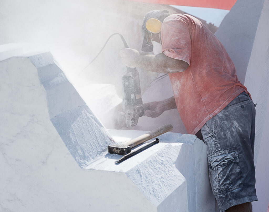 A worker, wearing protective gear, using a grinder on white stone which is producing a significant amount of dust.