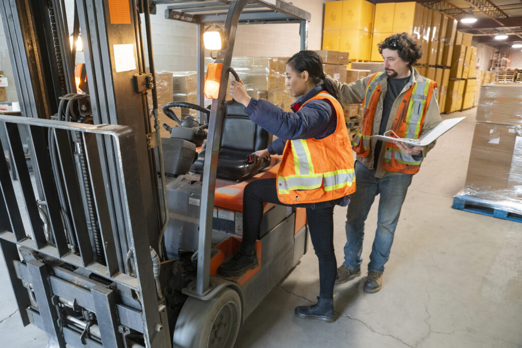 An industrial warehouse workplace safety topic.  A safety supervisor or manager training a new employee on forklift safety.  The trainer is holding a clipboard and explaining proper mounting of the forklift, with three points of contact.